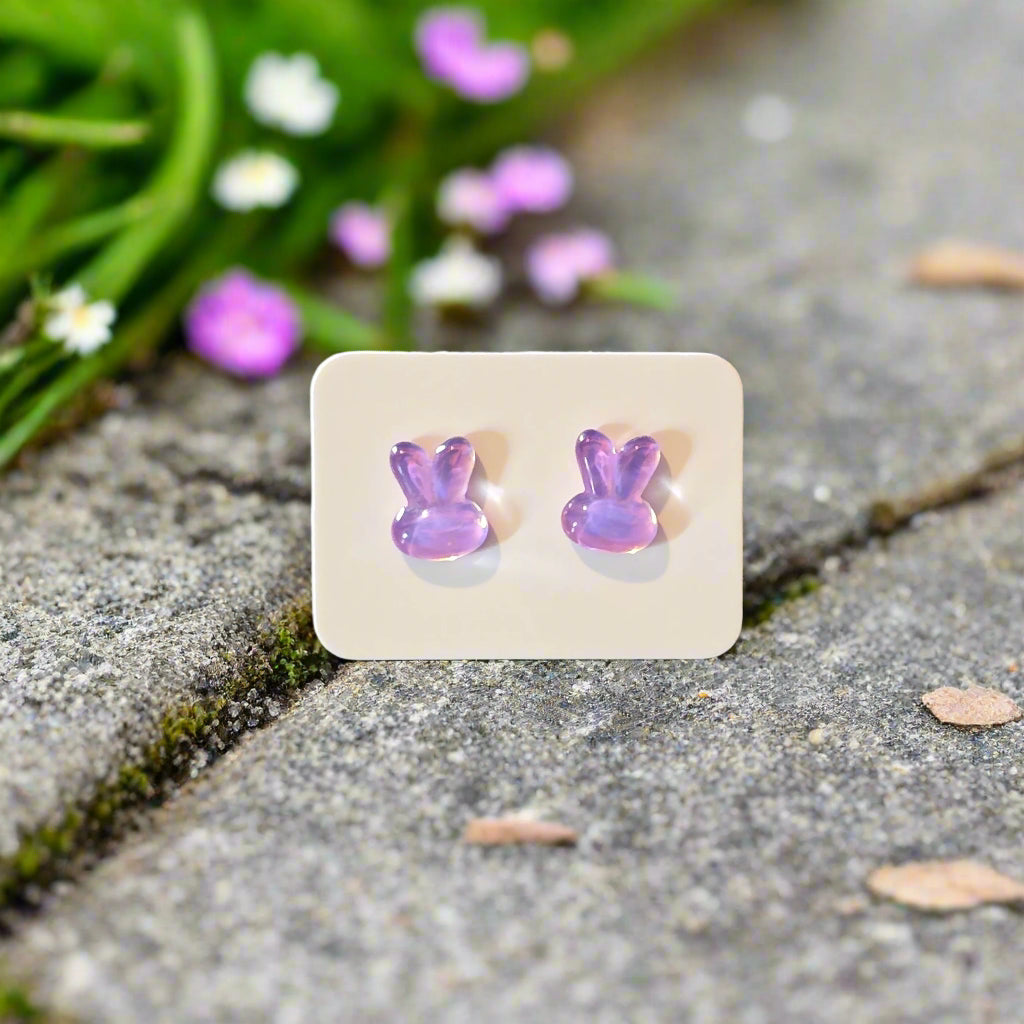 Purple bunny-shaped earrings on a beige card against an outdoor background